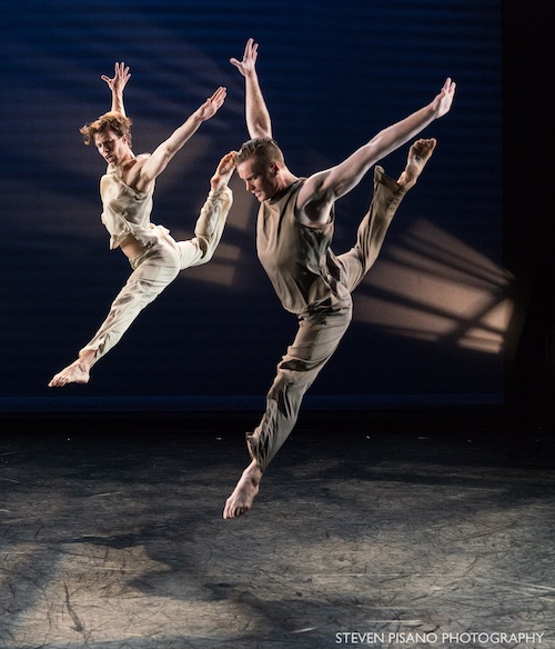 Two dancers leap into the air. One leg bends behind them while other extends in front. Their arms are above their head.